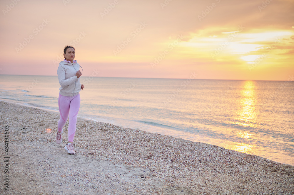 Full length attractive athletic woman doing morning jog along the coastline of the sea against the backdrop of a beautiful sunrise