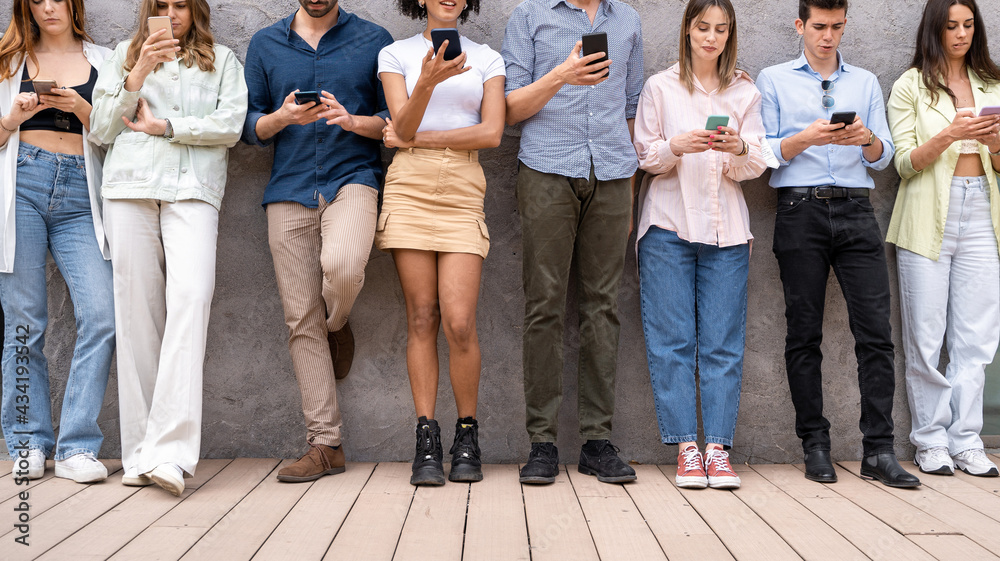 Group of young social network addicts people using smartphones browsing online standing against a wall