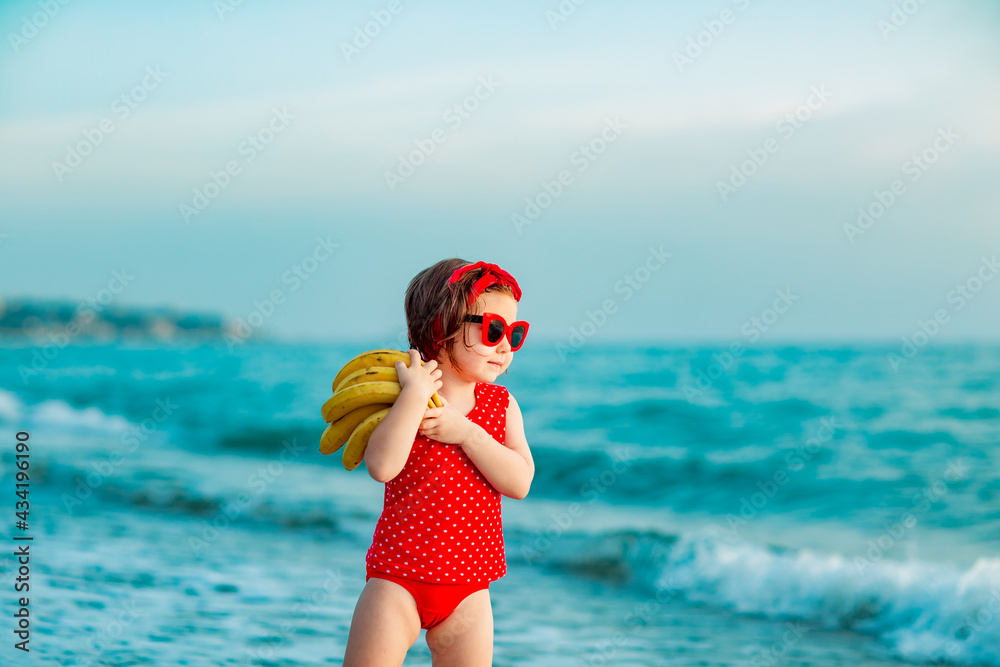 a little girl in a red swimsuit and sunglasses, stands on the beach, and holds ripe bananas in her hands
