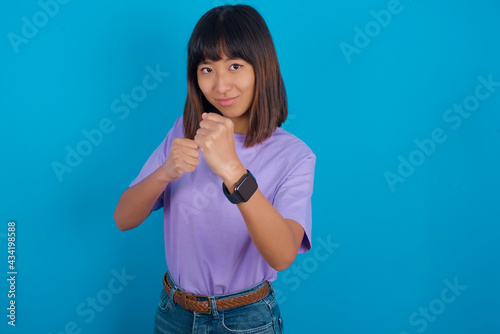 young beautiful asian woman wearing purple t-shirt against blue wall Ready to fight with fist defense gesture, angry and upset face, afraid of problem.