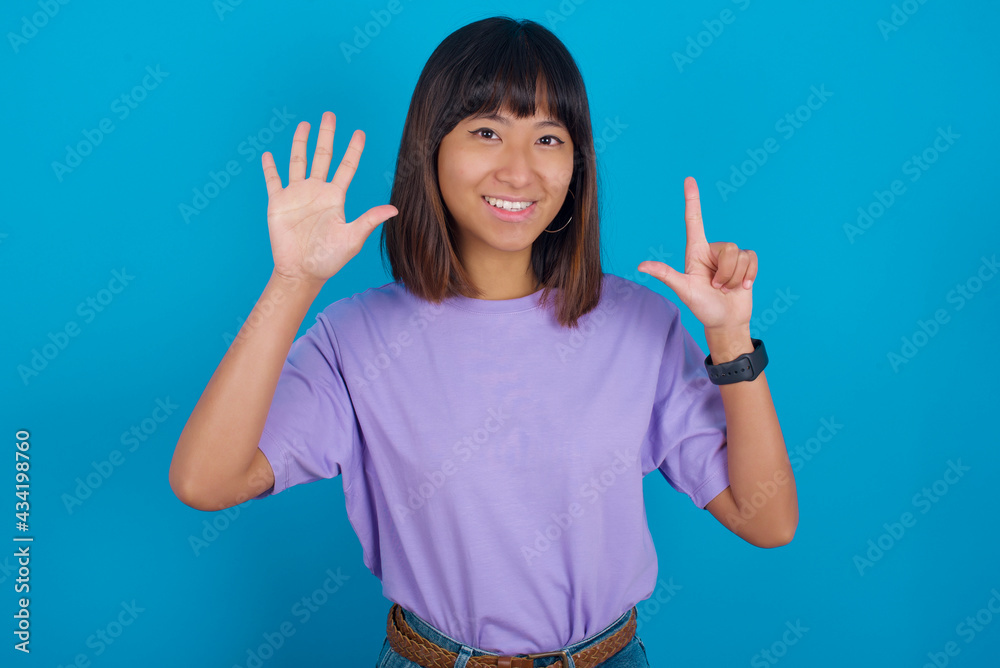 young beautiful asian woman wearing purple t-shirt against blue wall showing and pointing up with fingers number seven while smiling confident and happy. young beautiful asian woman wearing purple t-shirt against blue wall showing and pointing up with fingers number seven while smiling confident and happy.