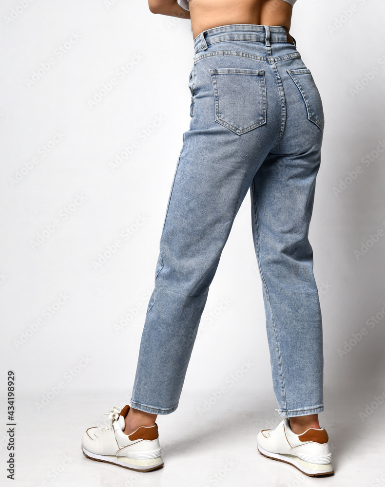 Unknown young skinny woman dressed in stylish blue jeans and white sneakers. Girl poses on a white background showing off pants and a perfect figure. Concept of a new collection of denim clothing. Unknown young skinny woman dressed in stylish blue jeans and white sneakers. Girl poses on a white background showing off pants and a perfect figure. Concept of a new collection of denim clothing.