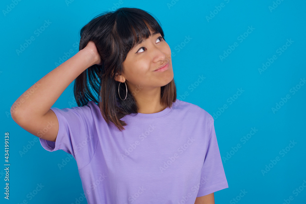 young beautiful asian woman wearing purple t-shirt against blue wall confuse and wonder about question. Uncertain with doubt, thinking with hand on head. Pensive concept. young beautiful asian woman wearing purple t-shirt against blue wall confuse and wonder about question. Uncertain with doubt, thinking with hand on head. Pensive concept.