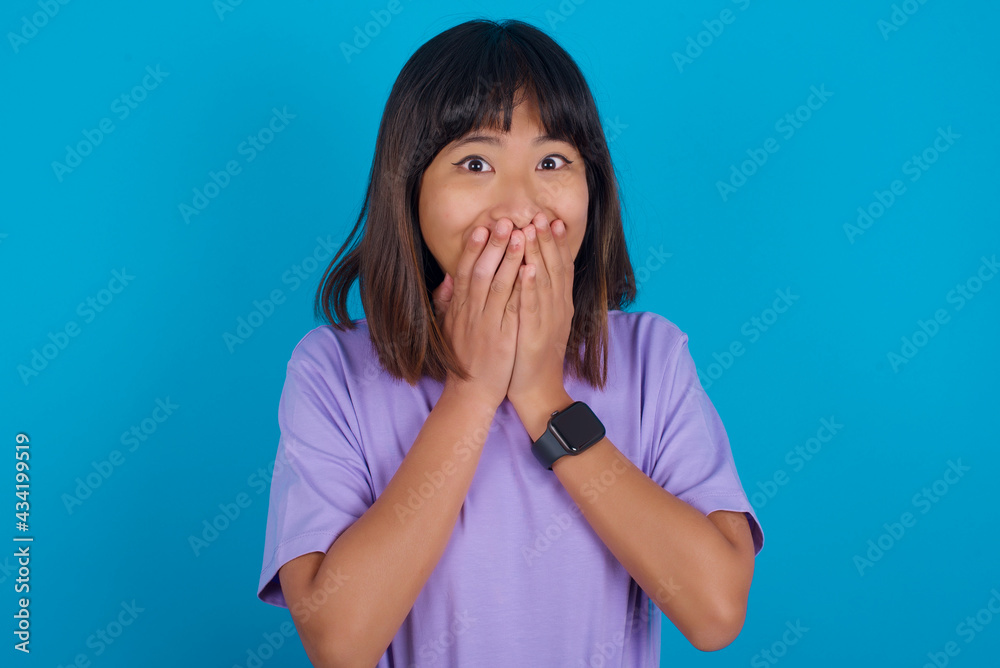 Vivacious young beautiful asian woman wearing purple t-shirt against blue wall, giggles joyfully, covers mouth, has natural laughter, hears positive story or funny anecdote Vivacious young beautiful asian woman wearing purple t-shirt against blue wall, giggles joyfully, covers mouth, has natural laughter, hears positive story or funny anecdote