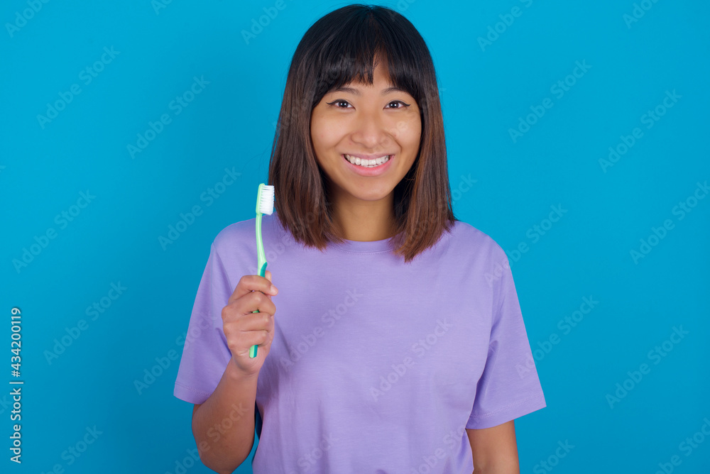 young beautiful asian woman wearing purple t-shirt against blue wall holding a toothbrush and smiling. Dental healthcare concept. young beautiful asian woman wearing purple t-shirt against blue wall holding a toothbrush and smiling. Dental healthcare concept.
