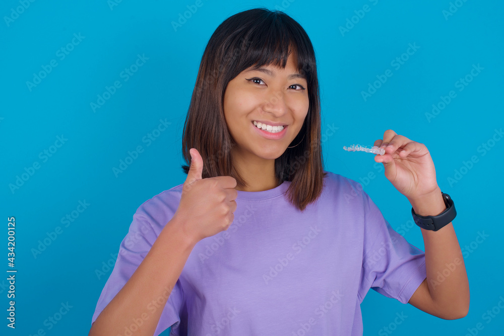 young beautiful asian woman wearing purple t-shirt against blue wall holding an invisible braces aligner and rising thumb up, recommending this new treatment. Dental healthcare concept. young beautiful asian woman wearing purple t-shirt against blue wall holding an invisible braces aligner and rising thumb up, recommending this new treatment. Dental healthcare concept.