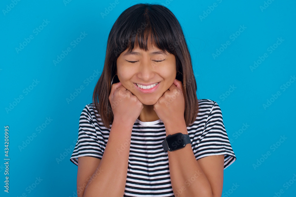 Cheerful young beautiful asian woman wearing stripped t-shirt against blue wall has shy satisfied expression, smiles broadly, shows white teeth, People emotions Cheerful young beautiful asian woman wearing stripped t-shirt against blue wall has shy satisfied expression, smiles broadly, shows white teeth, People emotions