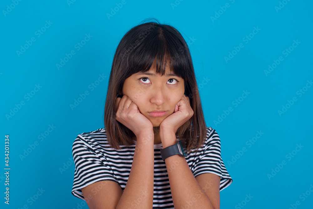 Portrait of sad young beautiful asian woman wearing stripped t-shirt against blue wall hands face look empty space Portrait of sad young beautiful asian woman wearing stripped t-shirt against blue wall hands face look empty space