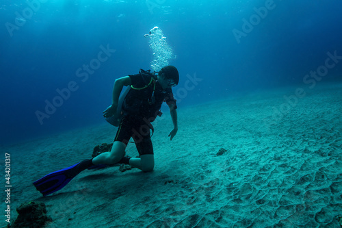 Young diver sits on sandy bottom on knees and carefully looking at something hiding under bottom surface - probably some fish or crab. His pose shows some danger - he moves hands back