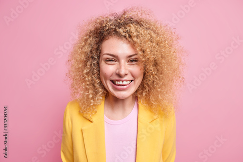 Portrait of attractive lady with curly bushy hair smiles toothily enjoys having nice day glad after successful deal wears formal costume isolated over pink background chuckles amused expresses joy