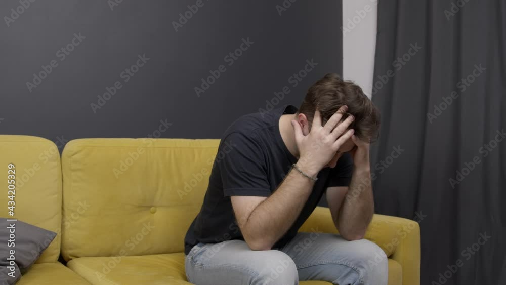 Young unshaven handsome guy sits at home on yellow sofa suffering from headache. Migraine, pain, stress, overwork, family problems. Painkillers, pain pills concept. Young unshaven handsome guy sits at home on yellow sofa suffering from headache. Migraine, pain, stress, overwork, family problems. Painkillers, pain pills concept.