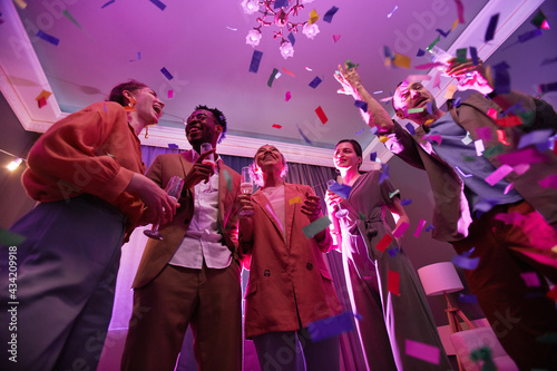 Low angle view at diverse group of young people standing under confetti shower while enjoying party with friends indoors