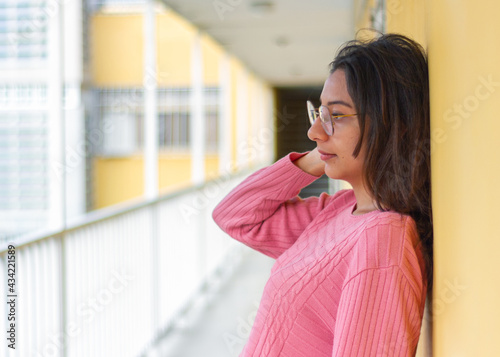 A pensive Latin woman standing on the hallway wearing glasses, with long dark hair, in a pink sweater