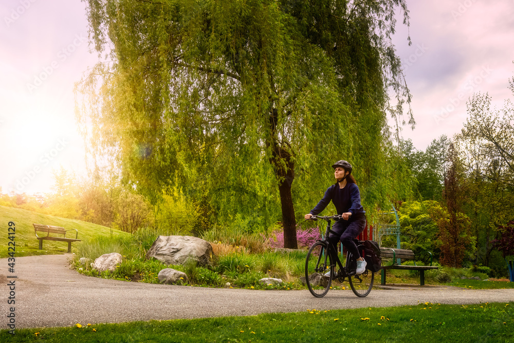 Adult Caucasian Woman Riding a Bicycle on a path in a modern city park. Spring Evening. Taken in Hawthorne Rotary Park, Surrey, Vancouver, British Columbia, Canada.