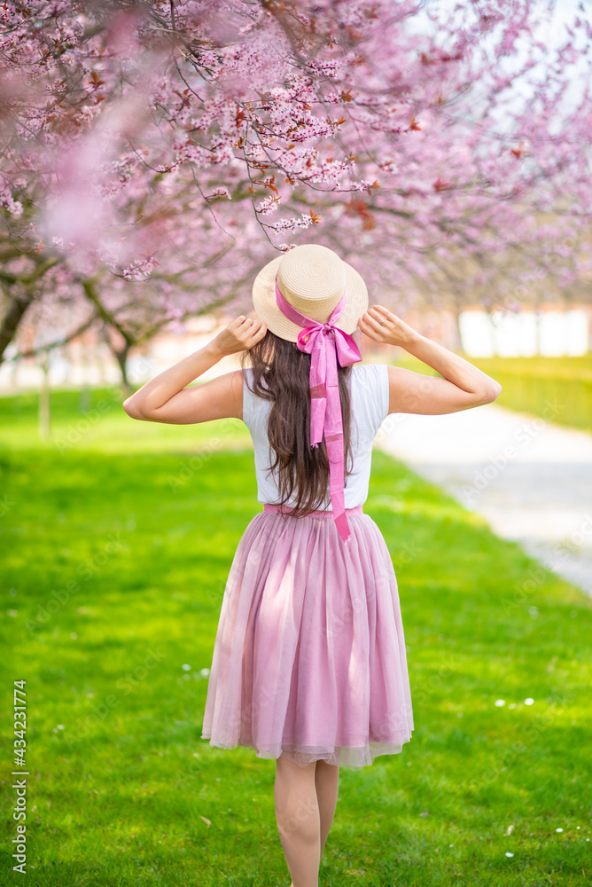 Beautiful woman in straw hat walking in a summer garden with blooming cherry trees. Girl wearing a pink long skirt