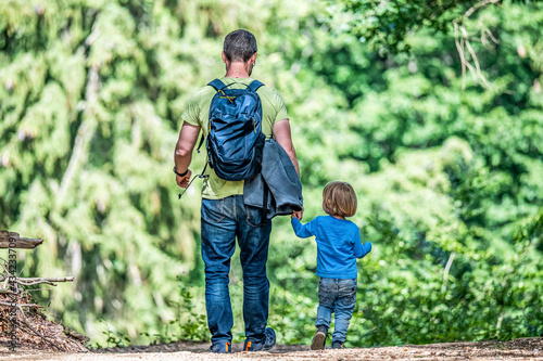 father holding hand of little son with backpack hiking in forest