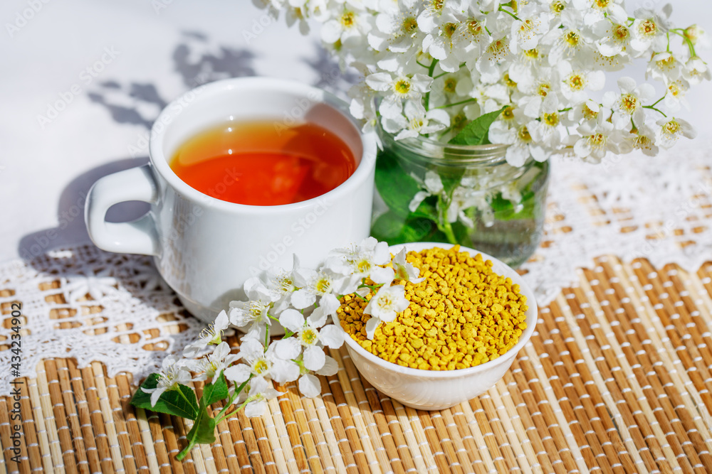 A bowl of pollen and aromatic herbal tea. Still life in a rustic style with a branch and flowers of a bird cherry and with a white knitted napkin. E A bowl of pollen and aromatic herbal tea. Still life in a rustic style with a branch and flowers of a bird cherry and with a white knitted napkin. E