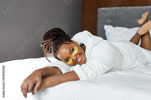 Modern cosmetology and anti-fatigue patches under eyes at home. Relaxed fat happy black woman apply gold patches under eyes, have rest on bed at weekends, beauty procedures after shower photo