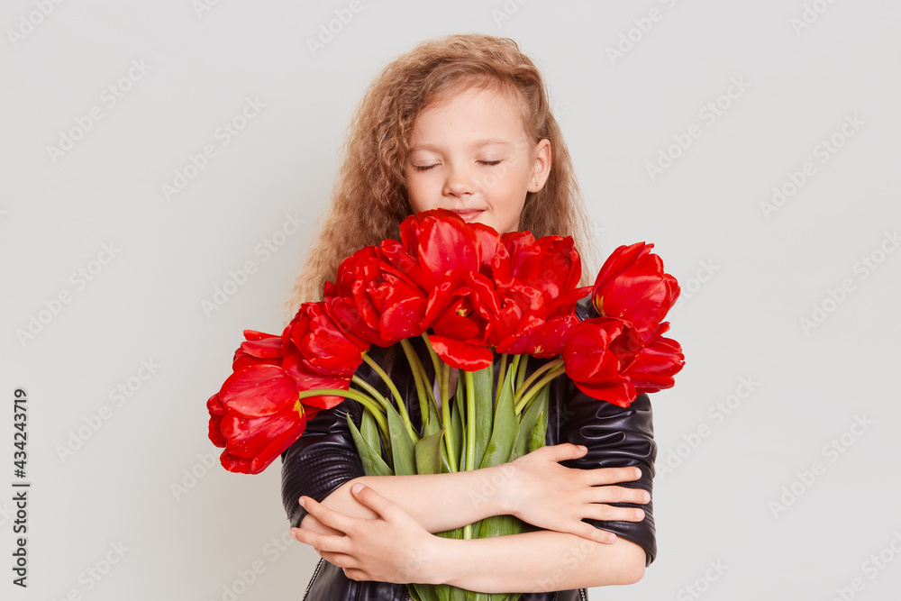 Happy dreamy little blonde girl embracing lots of red tulips and keeping eyes closed, enjoying beautiful present, wearing black jacket, isolated over gray background. Happy dreamy little blonde girl embracing lots of red tulips and keeping eyes closed, enjoying beautiful present, wearing black jacket, isolated over gray background.