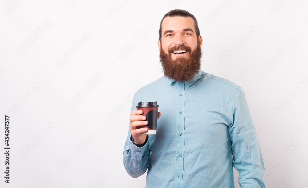 Naklejka premium A young bearded employee is smiling at the camera holding a cup of hot drink