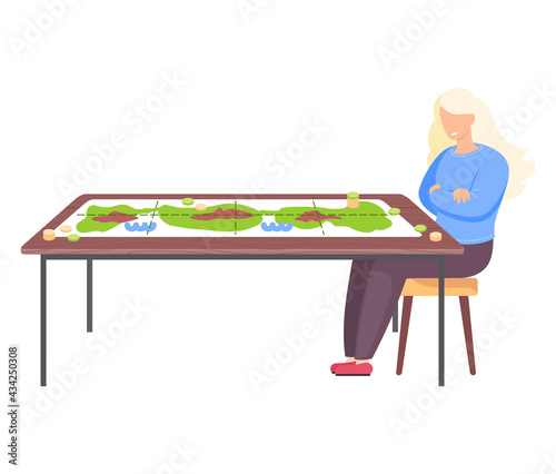 Girl sitting on chair next to table with colored board game and coins. Young woman has interesting hobby. Green map on table for playing with chips. Person resting and playing table game alone