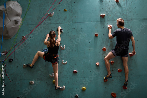 Rear view on couple climbers. Extreme indoor climbing. Strong woman and man practicing climbing on artificial rock wall in sports club. Slim sporty female and male training speed bouldering session.