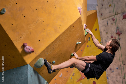 Young Man Climbs Up An Artificial Rock Wall,Sport And Fitness Concept, Side View. Fit Athlete Guy In Black Sportswear Engaged In Sport, Practice Strength