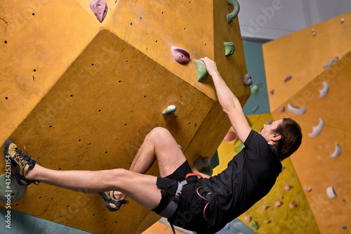 Male rock climber climbs artificial rock, man trains strength and endurance, overcoming the fear of heights, training indoors, alone, in sportive outfit. side view photo