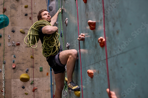 Caucasian strong sportsman climber moving up on steep rock, holding ropes on shoulders, climbing on artificial wall indoors, side view, copy space. Extreme sports and bouldering concept
