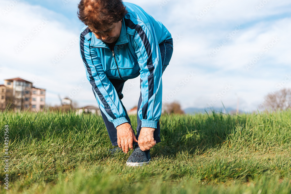 An elderly woman adjusts her sports shoes on her foot while standing on the grass. Pain in feet. The concept of an active lifestyle and health problems An elderly woman adjusts her sports shoes on her foot while standing on the grass. Pain in feet. The concept of an active lifestyle and health problems