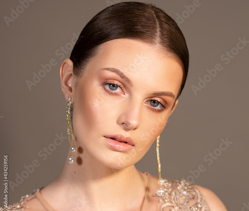 Portrait of an elegant woman. The hair is slicked back smoothly. In the ears are gold earrings with beads. Clean skin. Beige color. Look straight ahead. photo