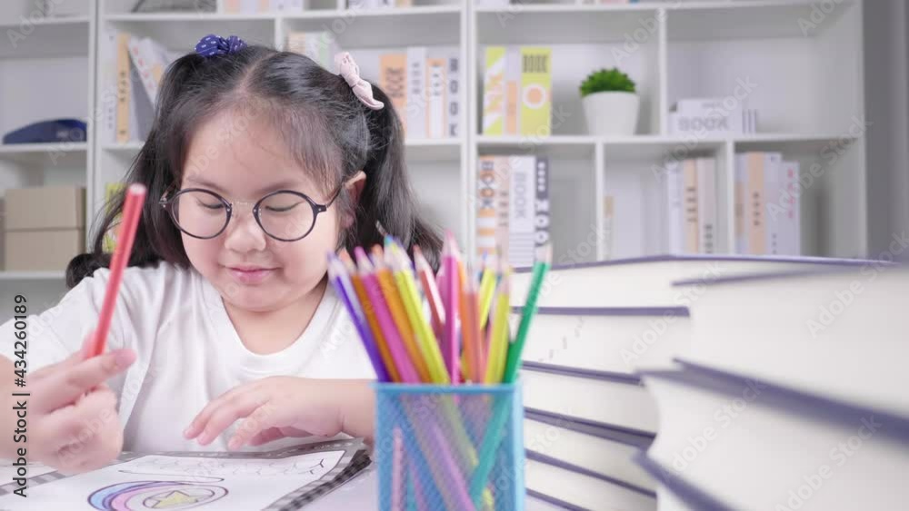 Asian girl wearing glasses is doing homework for school at the white deck, concentrating on her drawing and painting by coloured pencils in a cozy living room in her house after finishing school Asian girl wearing glasses is doing homework for school at the white deck, concentrating on her drawing and painting by coloured pencils in a cozy living room in her house after finishing school