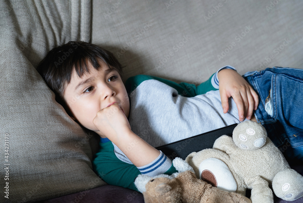 Candid shot of kid lying on sofa biting his finger nails while watching ...