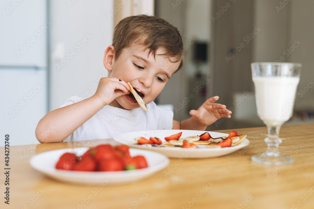 Cute toddler boy having breakfast with puncakes with berries and glass of milk in bright kitchen at home