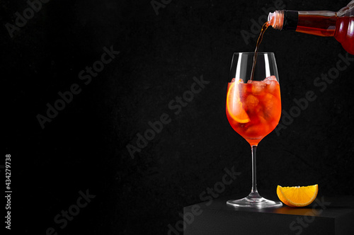 Pouring Aperol Spritz Cocktail in wine glass with ice and orange slice on dark background. Long fizzy drink