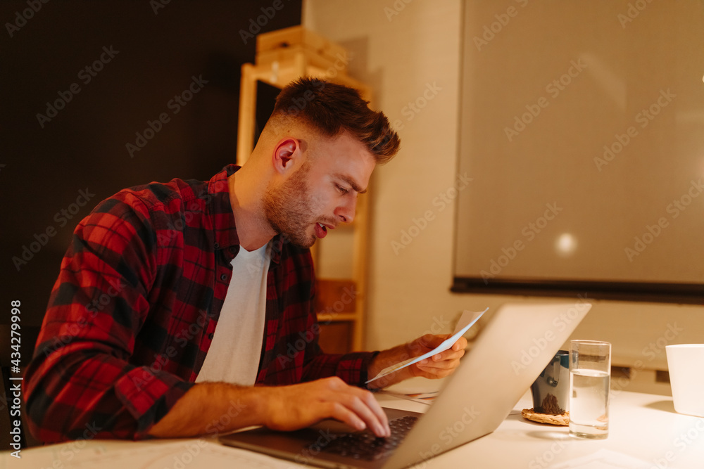 a young boy pays bills using a laptop. A young businessman works overtime from home.