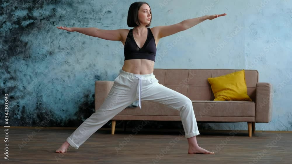 Young yogi woman practicing yoga, standing in Warrior two exercise, Virabhadrasana II pose in her living room, working out wearing sportswear bra and pants, full length. Weight loss concept . Young yogi woman practicing yoga, standing in Warrior two exercise, Virabhadrasana II pose in her living room, working out wearing sportswear bra and pants, full length. Weight loss concept .