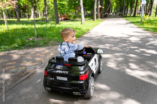 Back view of cute little caucasain blond toddler boy enjoy having fun riding electric powered police toy car by asphalt path road city park at summer day. Happy child playing rc vehicle outdoors