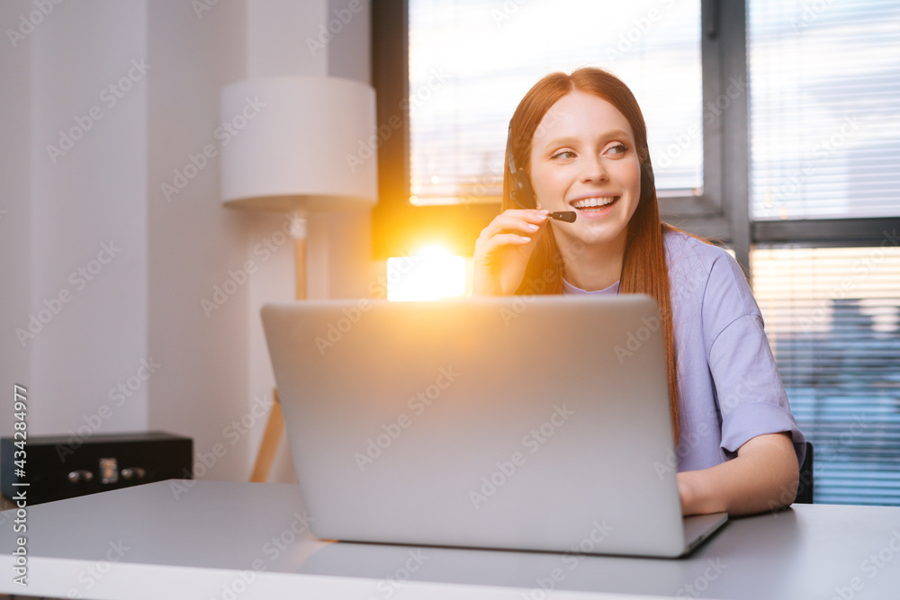 Close-up face of smiling young woman operator using headset and laptop during customer support at home office. Young redhead female student communicating online by video call on background of window. Close-up face of smiling young woman operator using headset and laptop during customer support at home office. Young redhead female student communicating online by video call on background of window.