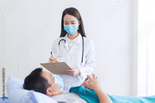 Asian female doctor is diagnosing and recording health of her patient who an asian man and stay on bed in hospital. Both of them wear the medical face mask to protect Covid 19 virus.