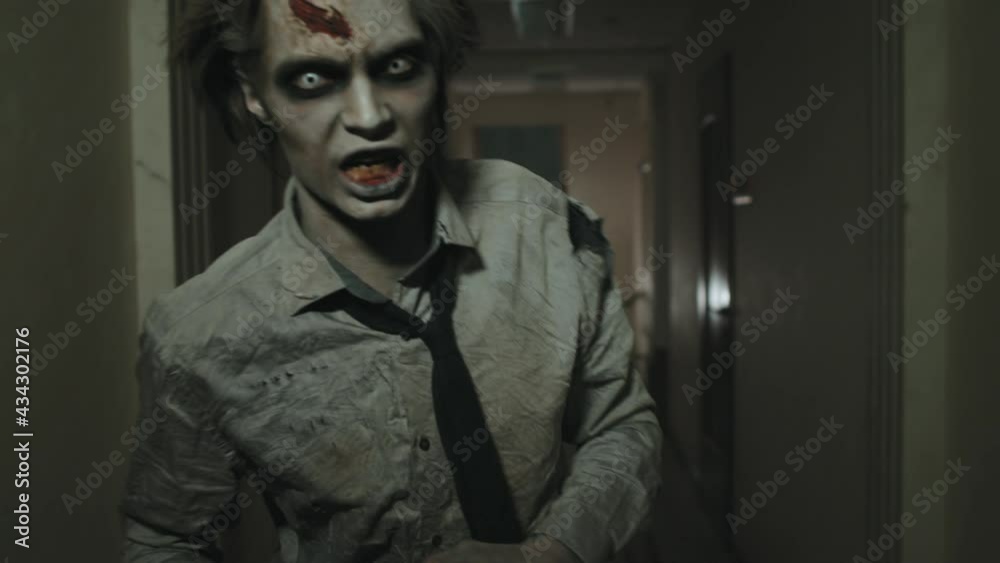 Tracking shot of creepy zombie man with SFX makeup and contact lenses running along dark hallway and following camera