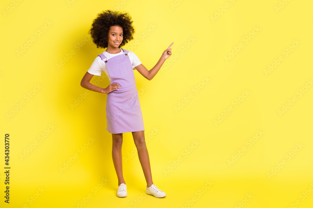 Full size photo of optimistic cute curly girl point empty space wear white t-shirt violet dress isolated on yellow color background Full size photo of optimistic cute curly girl point empty space wear white t-shirt violet dress isolated on yellow color background
