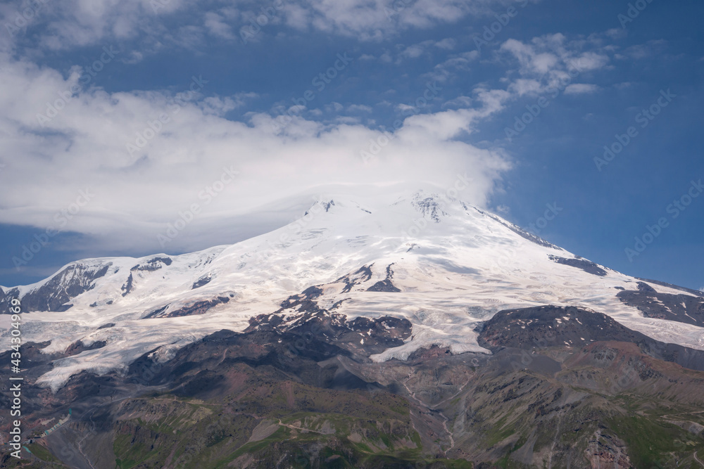 Volcano Elbrus. Its top is covered with clouds. They appeared in the afternoon. Landscape view in the south-east of mount from Cheget mount. Kabardino-Balkaria region, Russia.