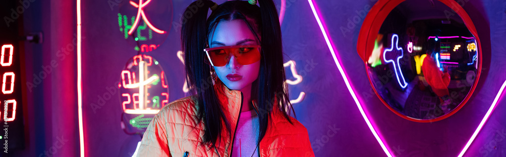 Obraz premium blue lighting on young asian woman in sunglasses posing near mirror and neon lighting, banner