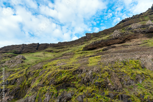 Low angle view of a steep grassy cliff against blue sky with clouds in Iceland in summer. Natural background.