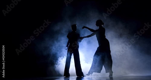 In the twilight on the stage, a ballroom couple is dancing masterly, They are illuminated by spotlights, smoke is around them. Only their silhouette is visible, they make fast and artistic movements