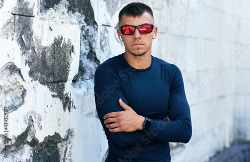 Sportsman taking a rest crossing with his arms outside. Portrait of an athlete man in blue sportswear and red sunglasses looking at he camera and posing against an urban concrete wall.