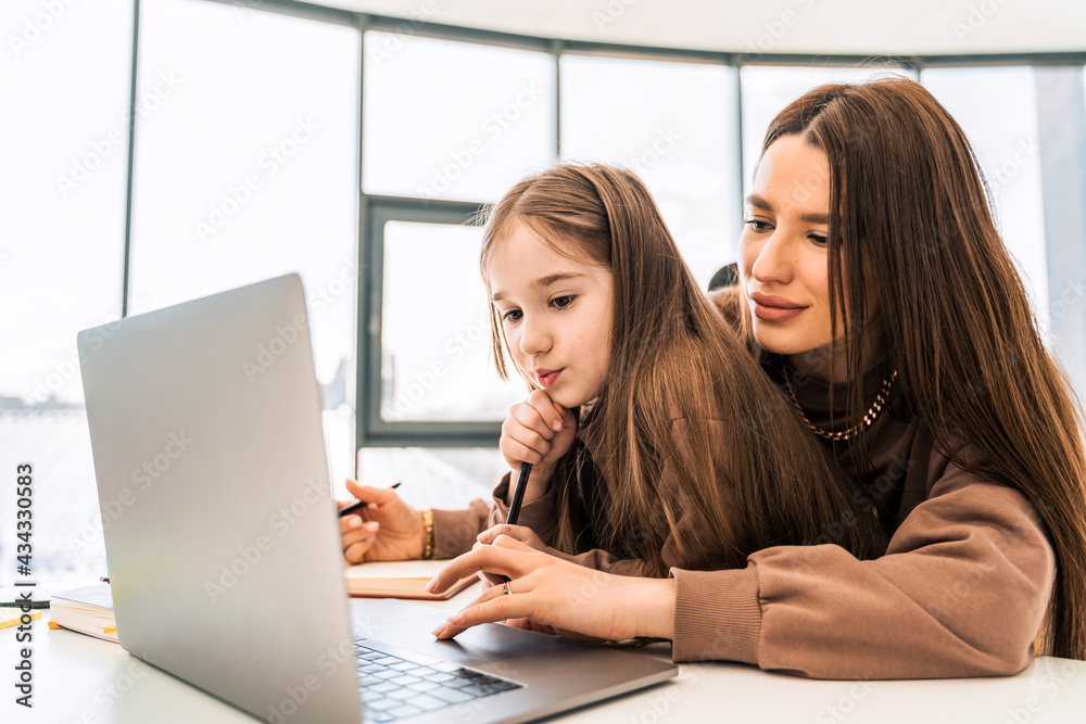 Mother and daughter are doing homework at the laptop. Little schoolgirl at the distance learning at home Mother and daughter are doing homework at the laptop. Little schoolgirl at the distance learning at home