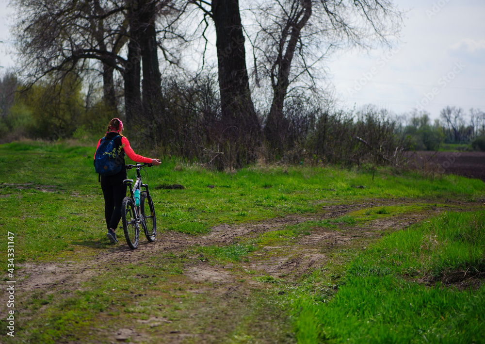 Fototapeta premium a girl with a bicycle walks a field trail. Woman riding a mountain bike in the forest. Hiker woman with backpack. one young woman riding a bike in nature. rolling the bike in his hands. back view.