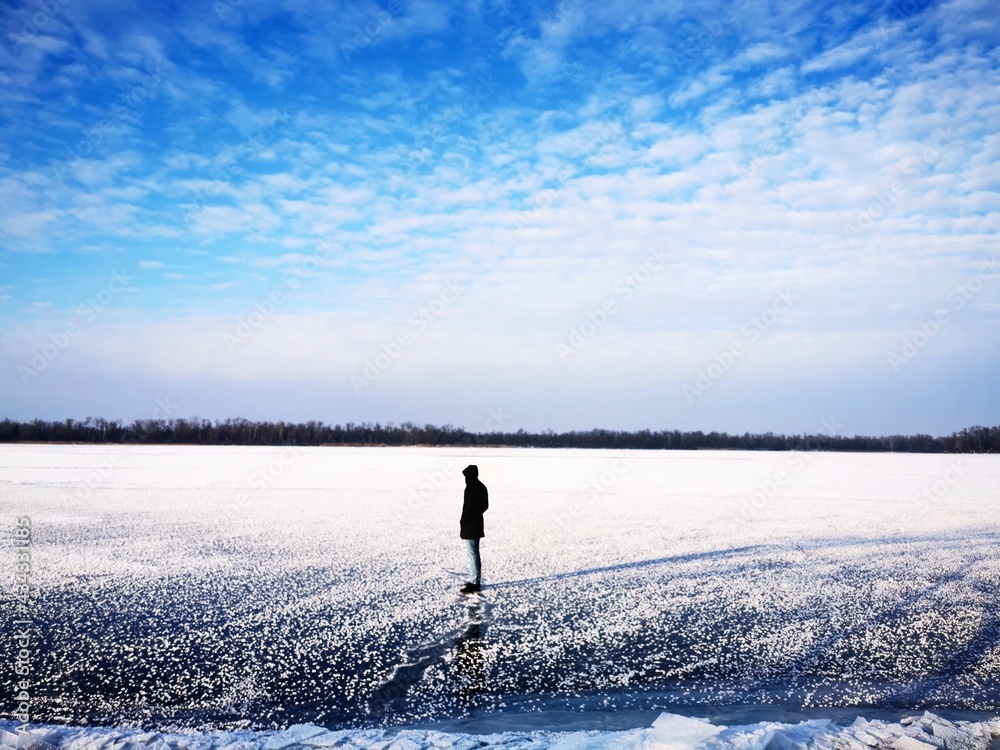  A silhouette of a lonely man in a dark coat stands on the ice of the river. Winter landscape photo